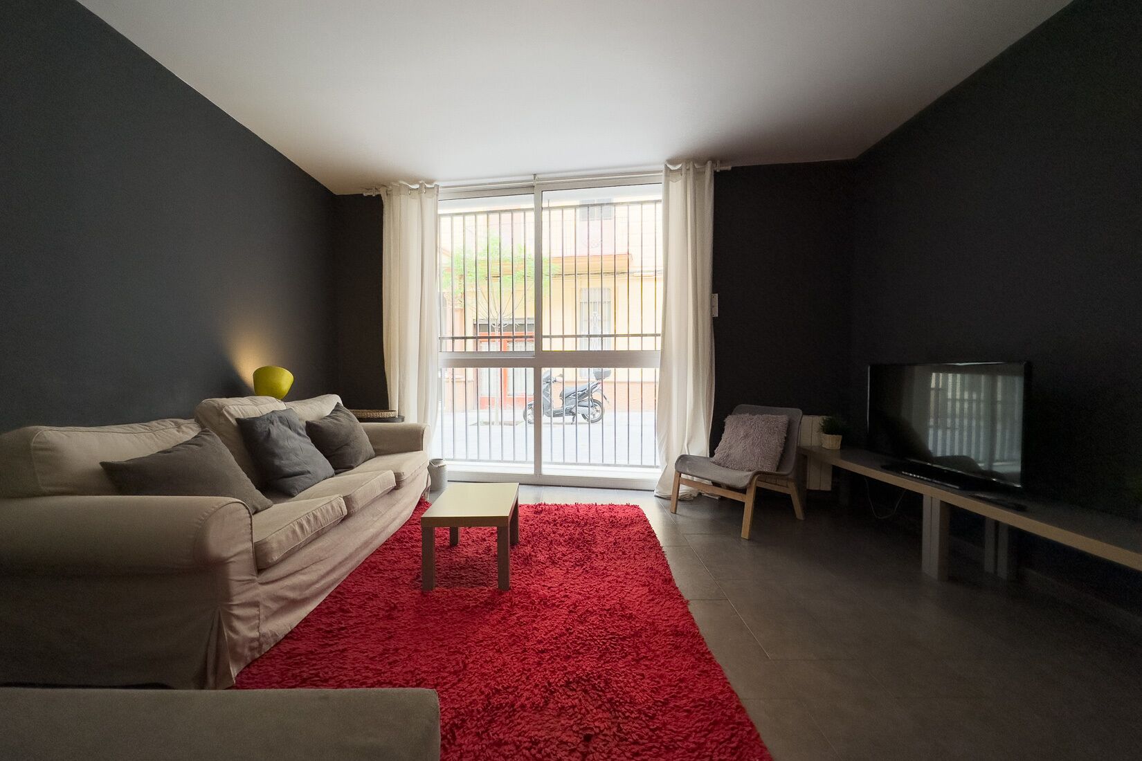 Ein Wohnzimmer mit einer Couch, einem roten Teppich, einem Fernseher und einem Fenster mit Blick ins Freie.