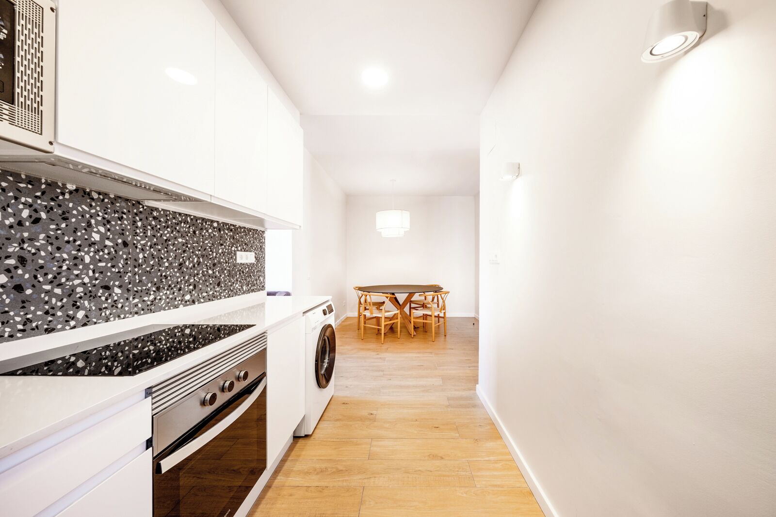 A compact kitchen with a washing machine, a dining area with a table and chairs, and a light fixture on the wall.