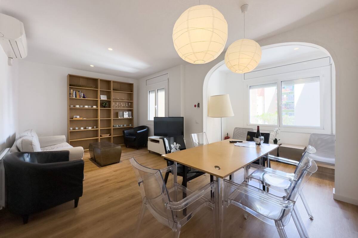 A modern living room with a wooden floor, a sofa, a coffee table, a TV, a bookshelf, and a dining area with a table and chairs.