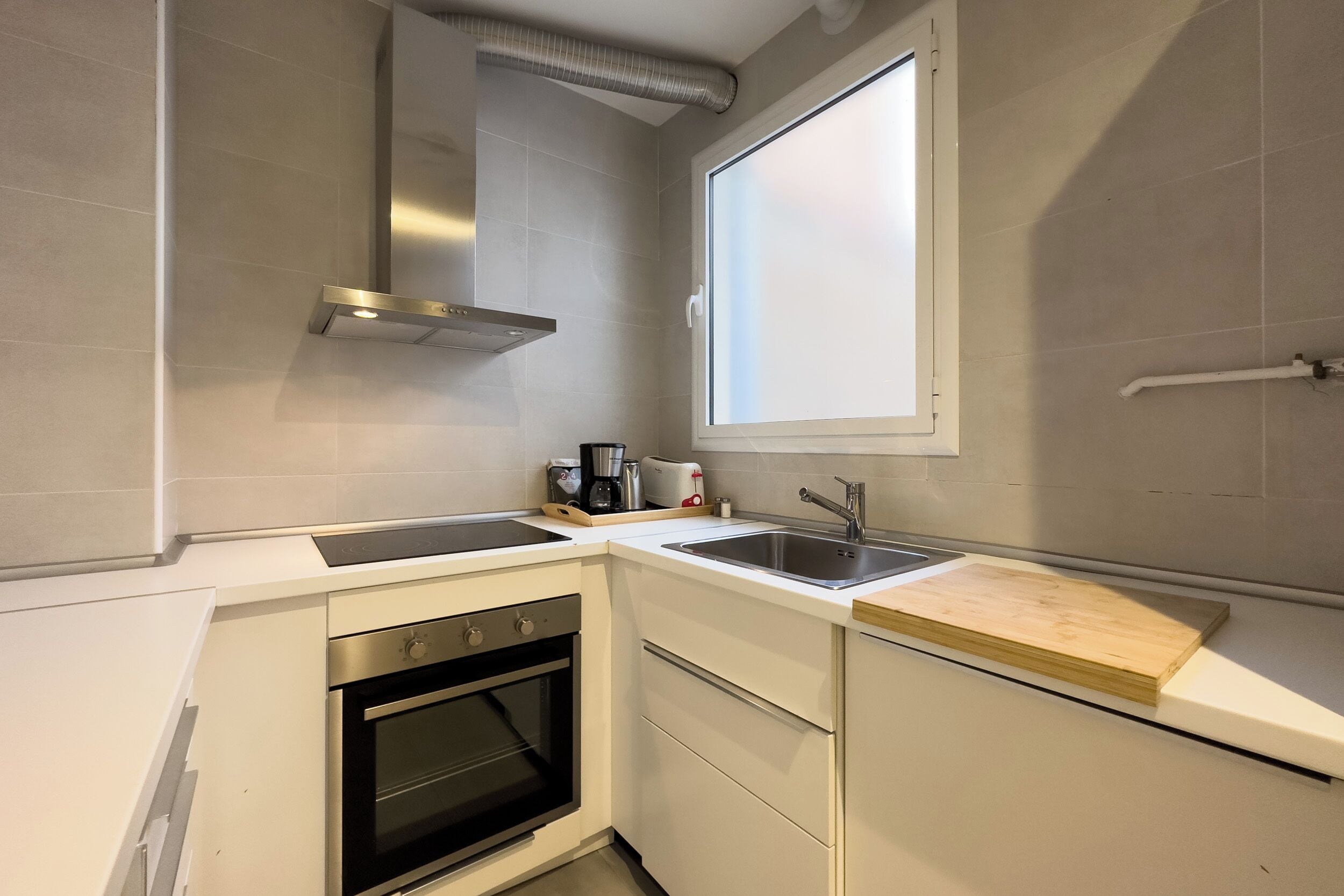 Una cocina compacta con armarios blancos, horno integrado, pileta de acero inoxidable y una ventana sobre la pileta.
