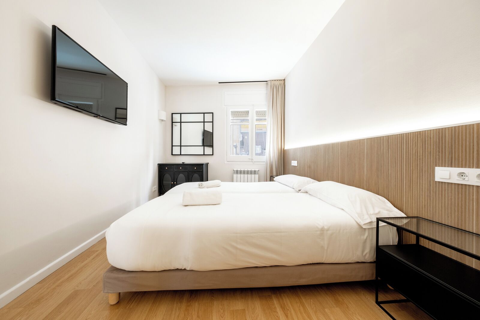 Una habitación de hotel moderna con una cama grande, un televisor de pantalla plana montado en la pared, un cabecero de madera y una mesita de noche pequeña.