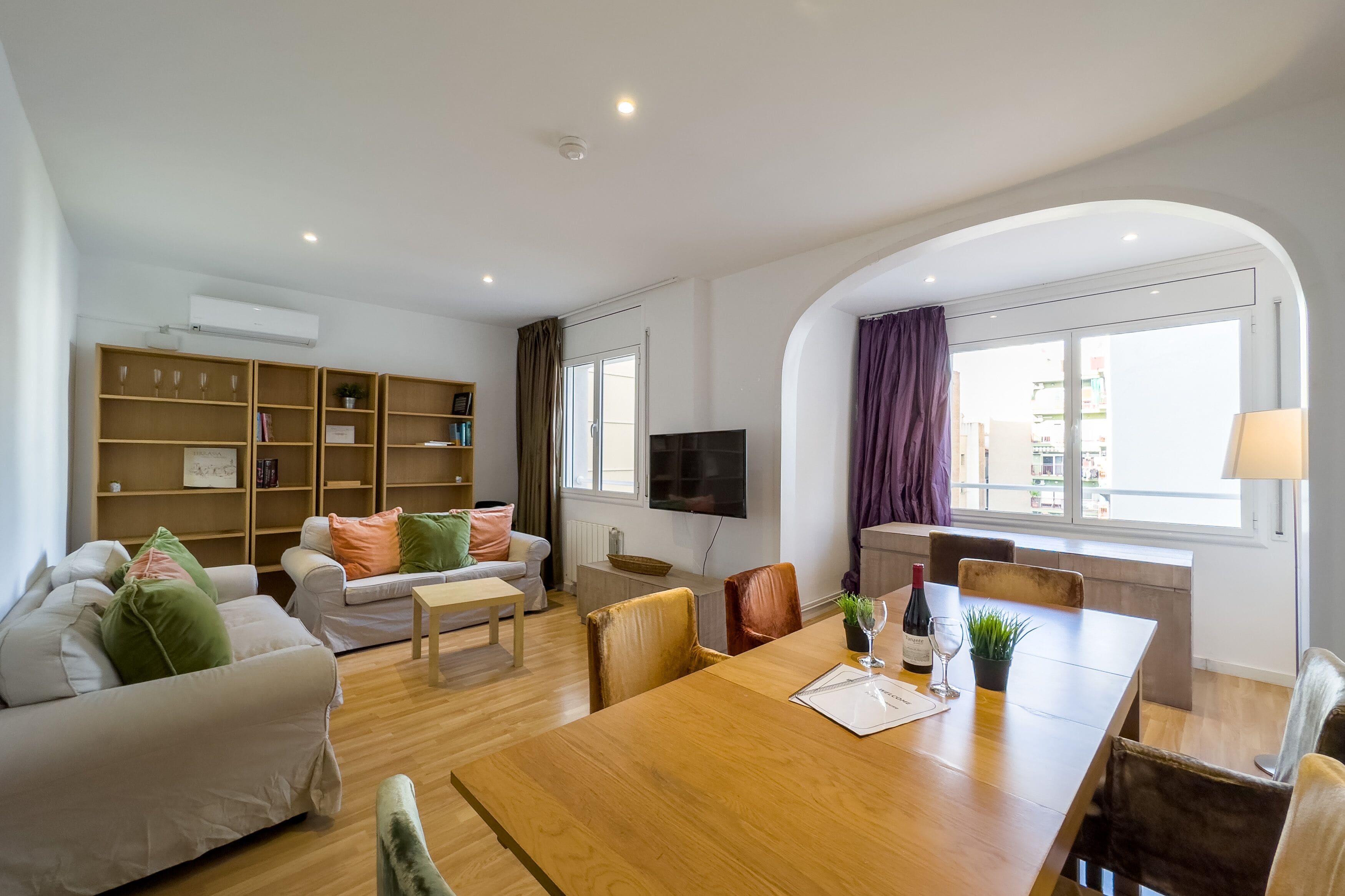 A modern living room with a wooden dining table, sofas, a TV, and a large window with curtains.