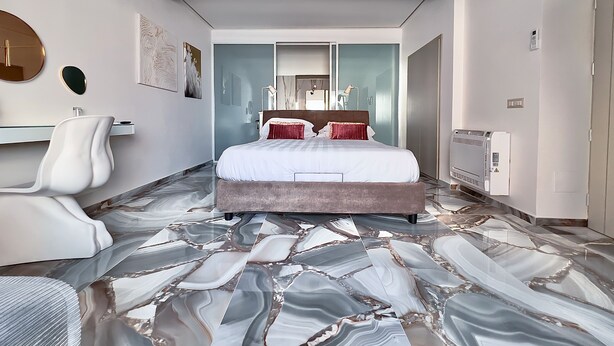 Een moderne hotelkamer met een marmeren vloer, een groot bed met rode en witte beddengoed, een witte fauteuil en een badkamer met spiegel en wastafel.
