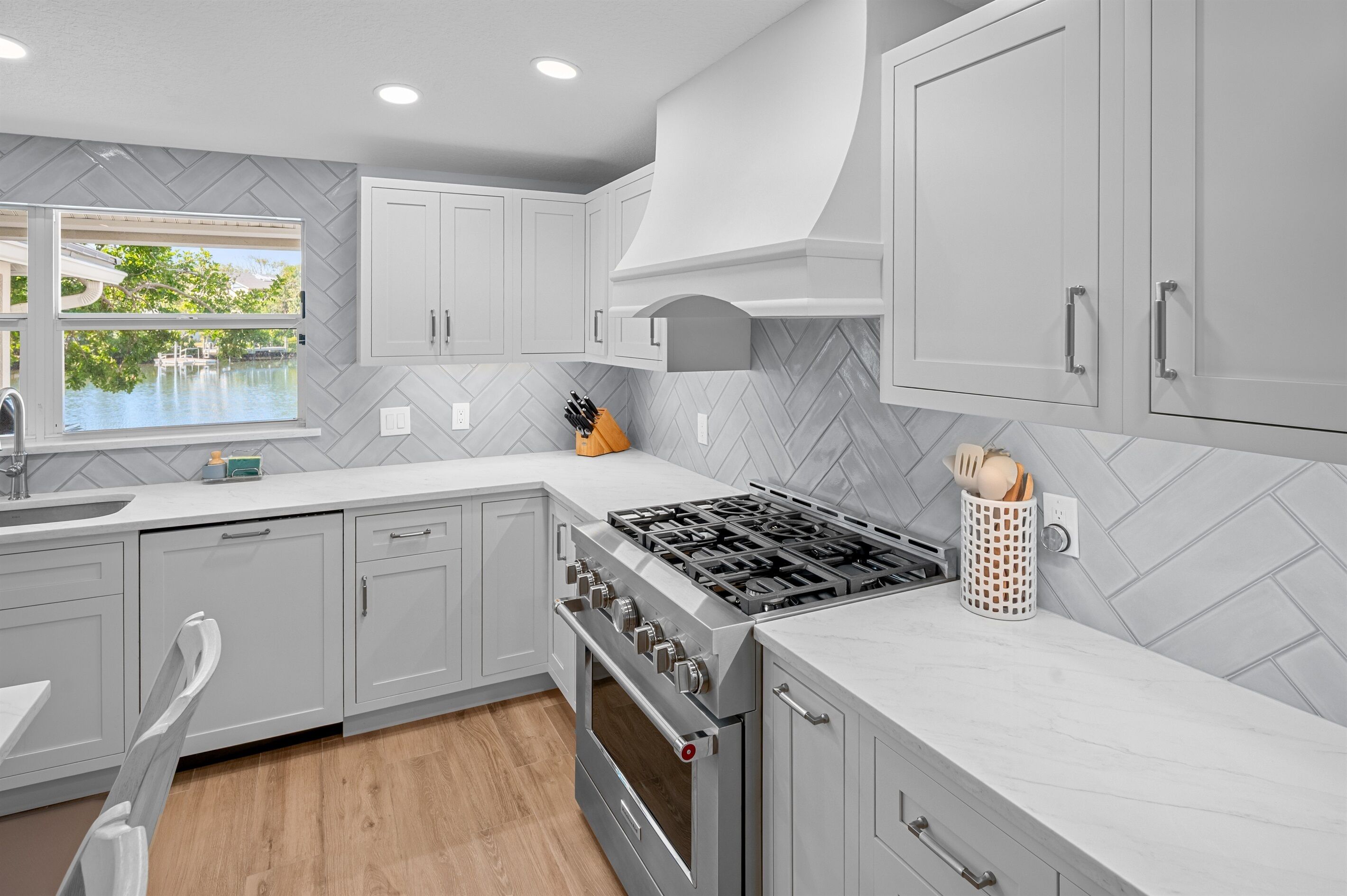 A modern kitchen with grey cabinetry, a herringbone-patterned backsplash, a gas stove, and a window overlooking a pool.