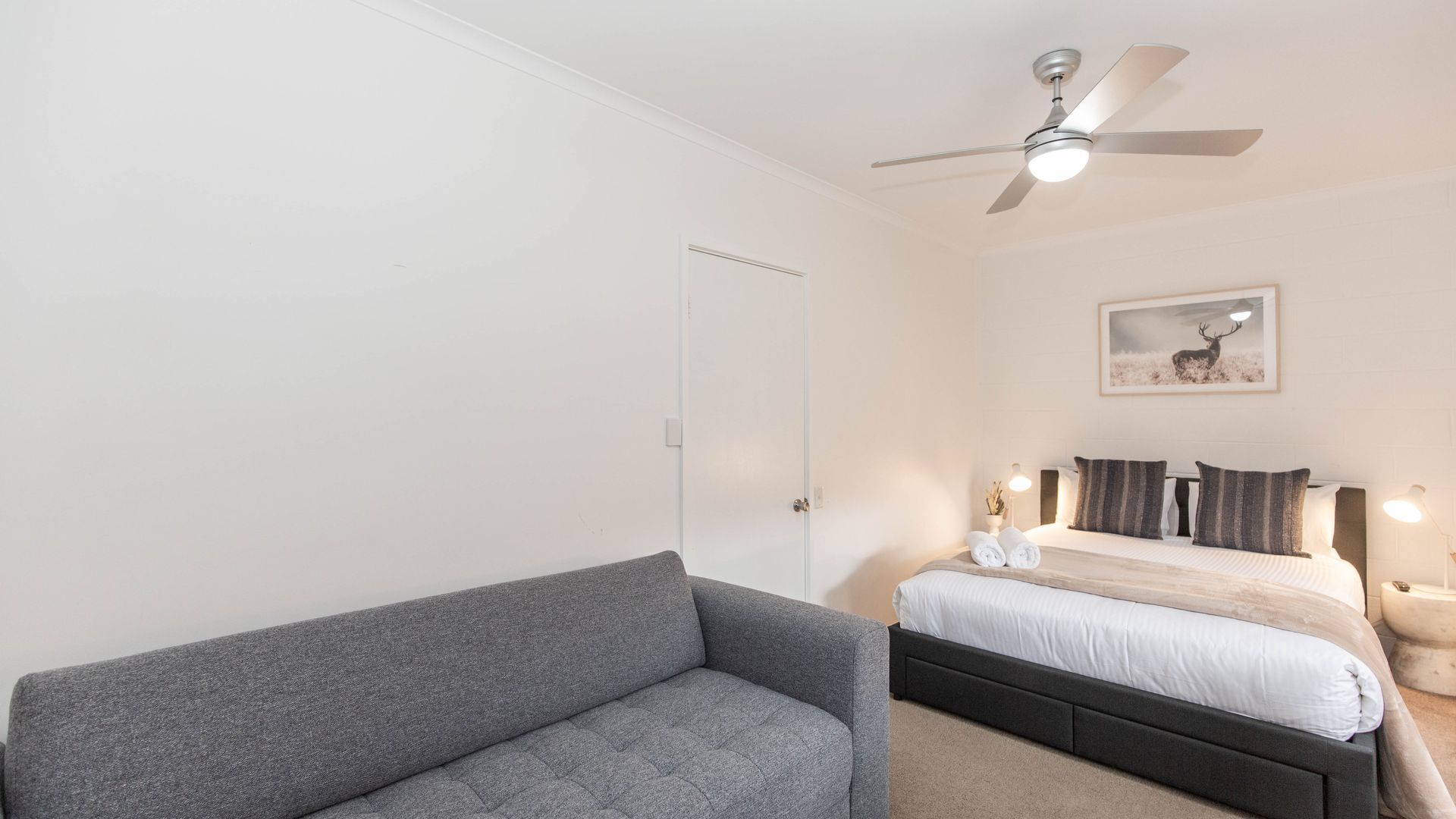 A bedroom with a bed, a grey sofa, a ceiling fan, and a framed picture on the wall.