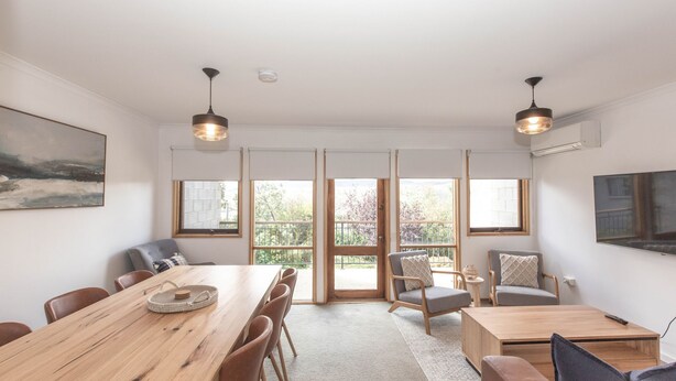 A spacious room with a large wooden table, multiple chairs, and a view of greenery through large windows.