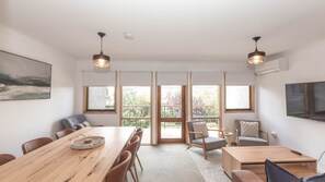 A spacious room with a large wooden table, multiple chairs, and a view of greenery through large windows. - Montebello 3 / 12 Cobbodah Street (Jindabyne)