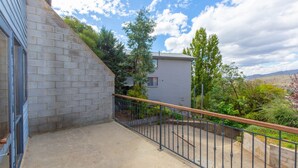 A balcony with a metal railing, a stone wall, and a view of a building with a window and trees in the background.