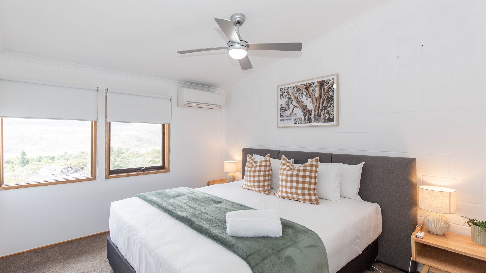 A bedroom with a bed, two windows, a ceiling fan, a wall-mounted air conditioner, and a framed picture.