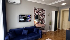 A modern living room with a blue sofa, a flat-screen TV, a floral wallpaper accent wall, a round mirror, and a wooden herringbone floor.