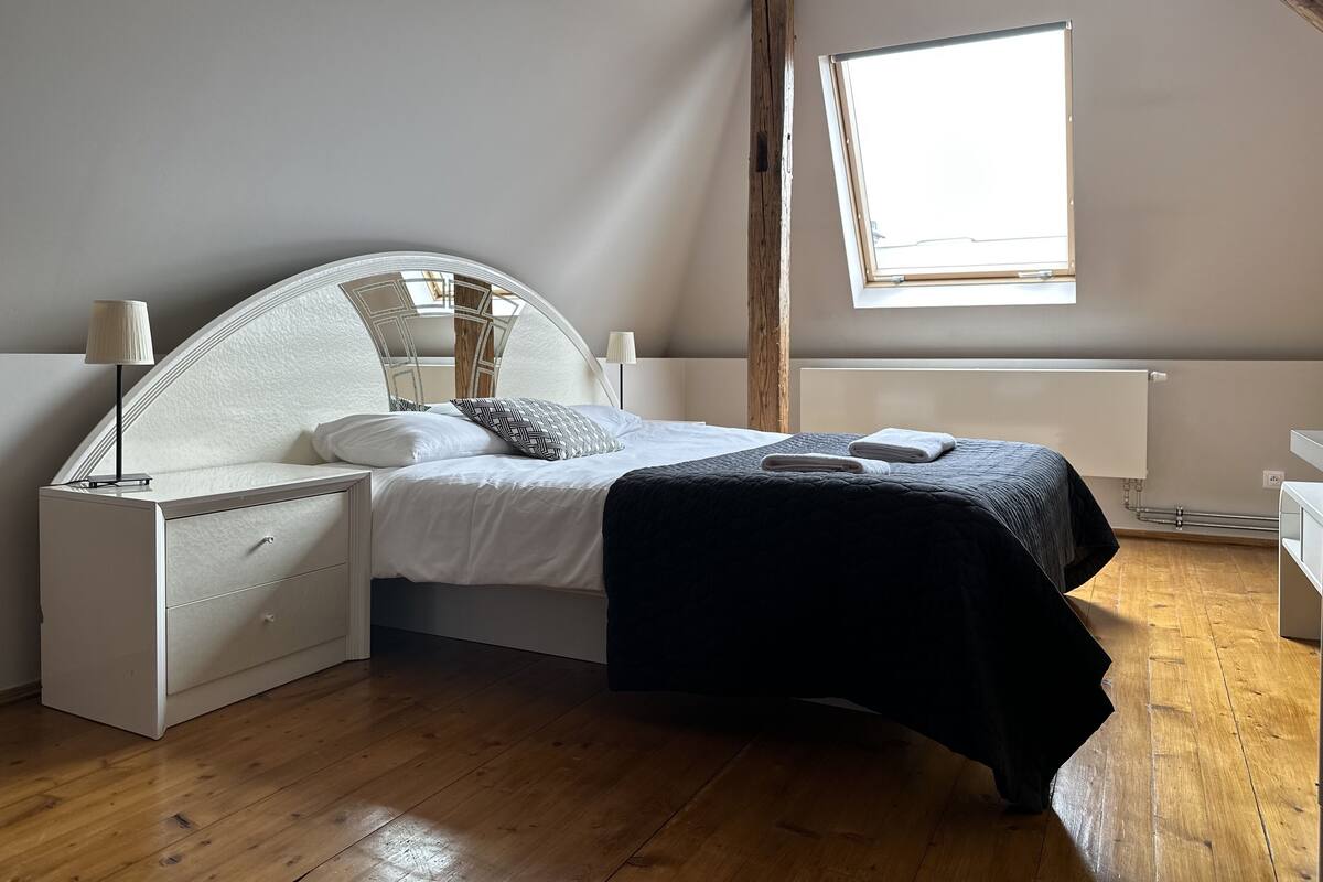 A bedroom with a curved headboard, a bed with white bedding and a black throw, wooden flooring, a window, and a radiator.
