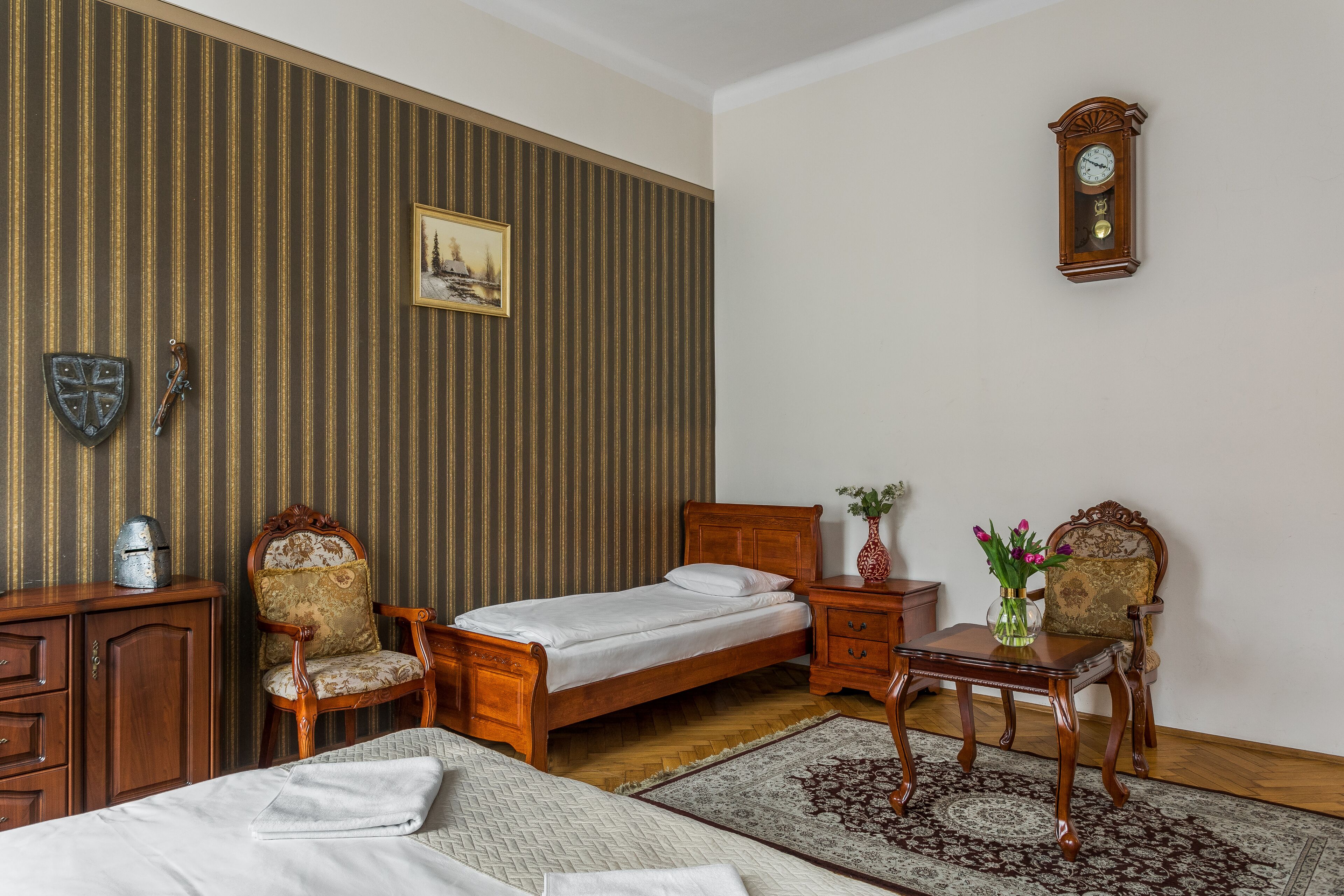 A bedroom with a wooden bed, a nightstand, a chair, a dresser, a clock on the wall, and a painting.