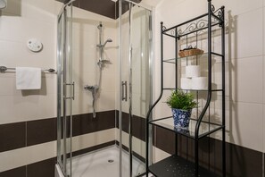 Ducha con cerramiento de vidrio, panel de control, perchero con toalla blanca y estante de metal negro con planta en maceta y artículos de aseo.