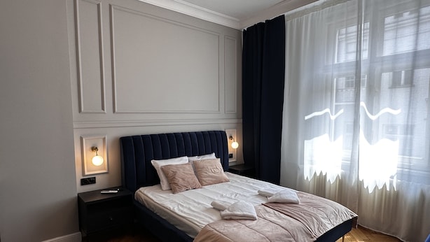 A bedroom with a large bed, two bedside tables with lamps, a window with sheer curtains, and a herringbone-patterned wooden floor.