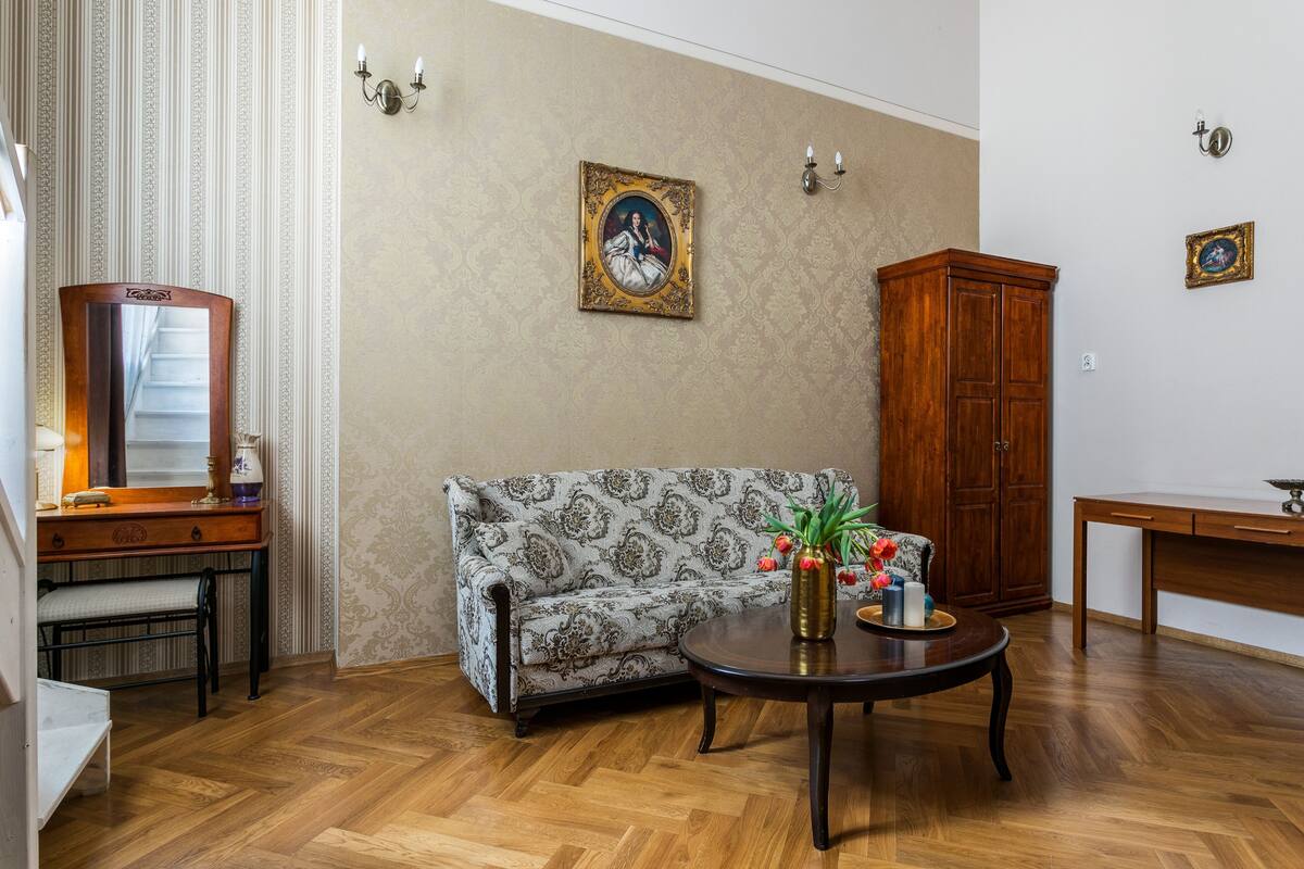 A room with a patterned sofa, a wooden cabinet, a round table with a vase and flowers, and a framed picture on the wall.