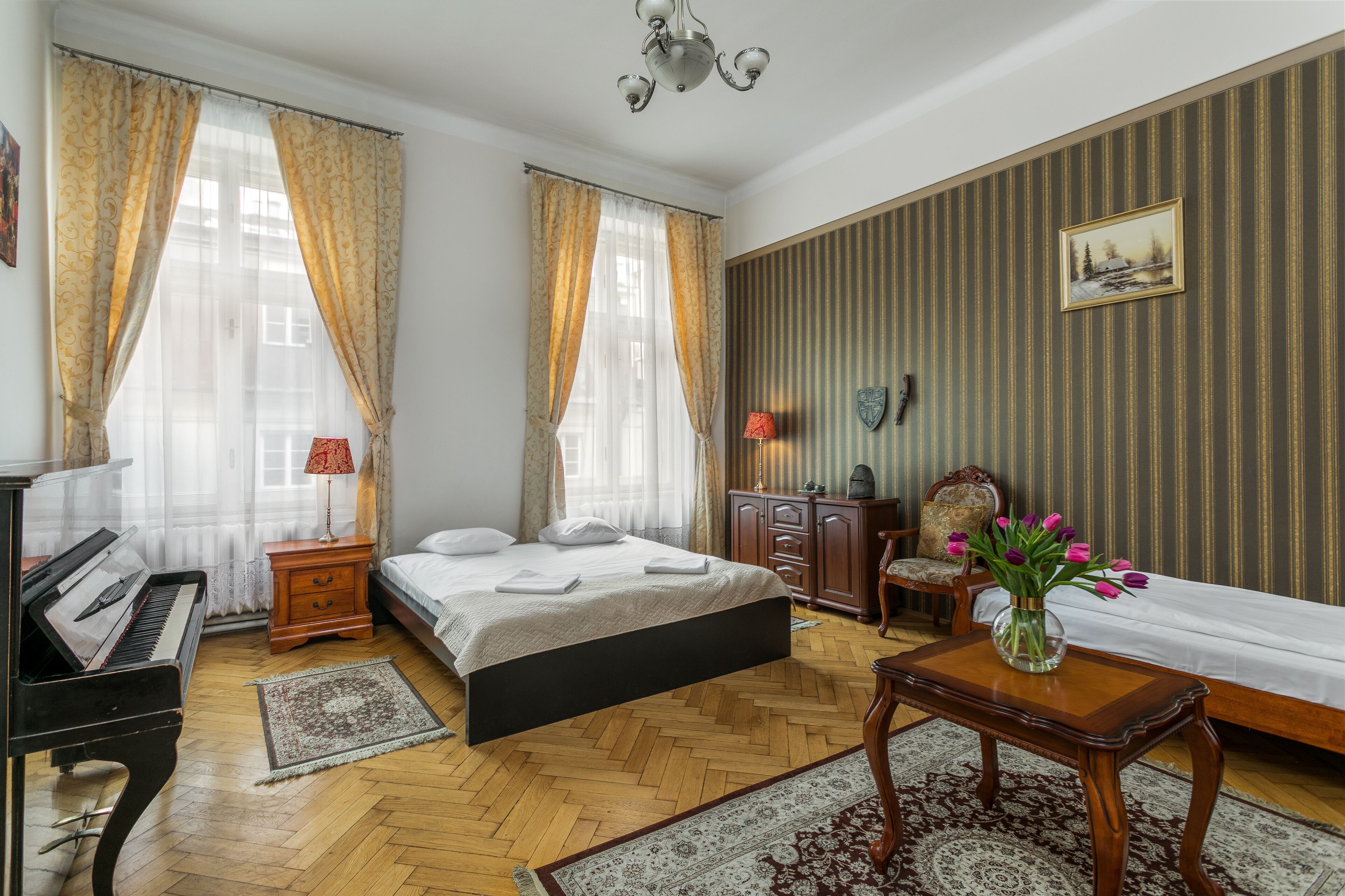 Una camera d'albergo con un pianoforte, un letto con lenzuola bianche, un comò di legno e un vaso con fiori.