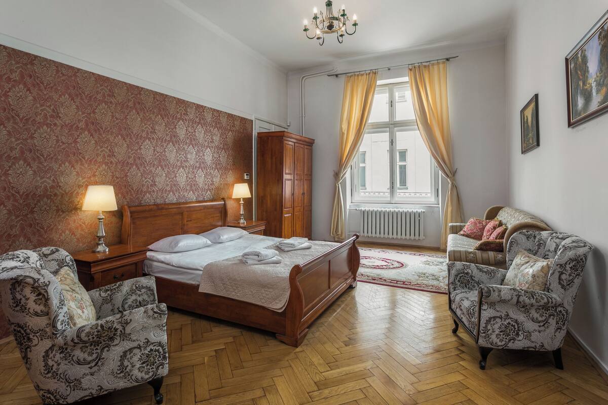 A bedroom with a wooden bed, two bedside tables with lamps, a wardrobe, patterned armchairs, a red patterned wall, and a window with curtains.