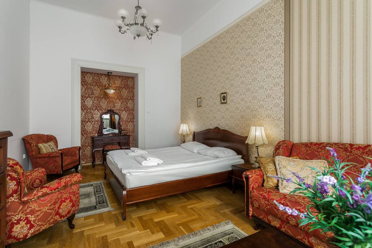 A hotel room with a wooden bed, red upholstered chairs, a wooden nightstand, and a chandelier.