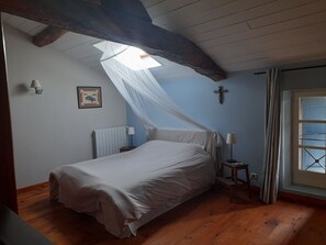 Une chambre à coucher comprenant un lit, une table de chevet, une lampe, une fenêtre avec des rideaux, un radiateur et un crucifix fixé au mur.