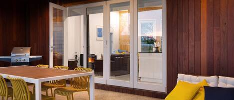 A cozy outdoor seating area with a wooden table and chairs, a barbecue grill, and a sliding glass door leading to a balcony with a sofa and cushions.