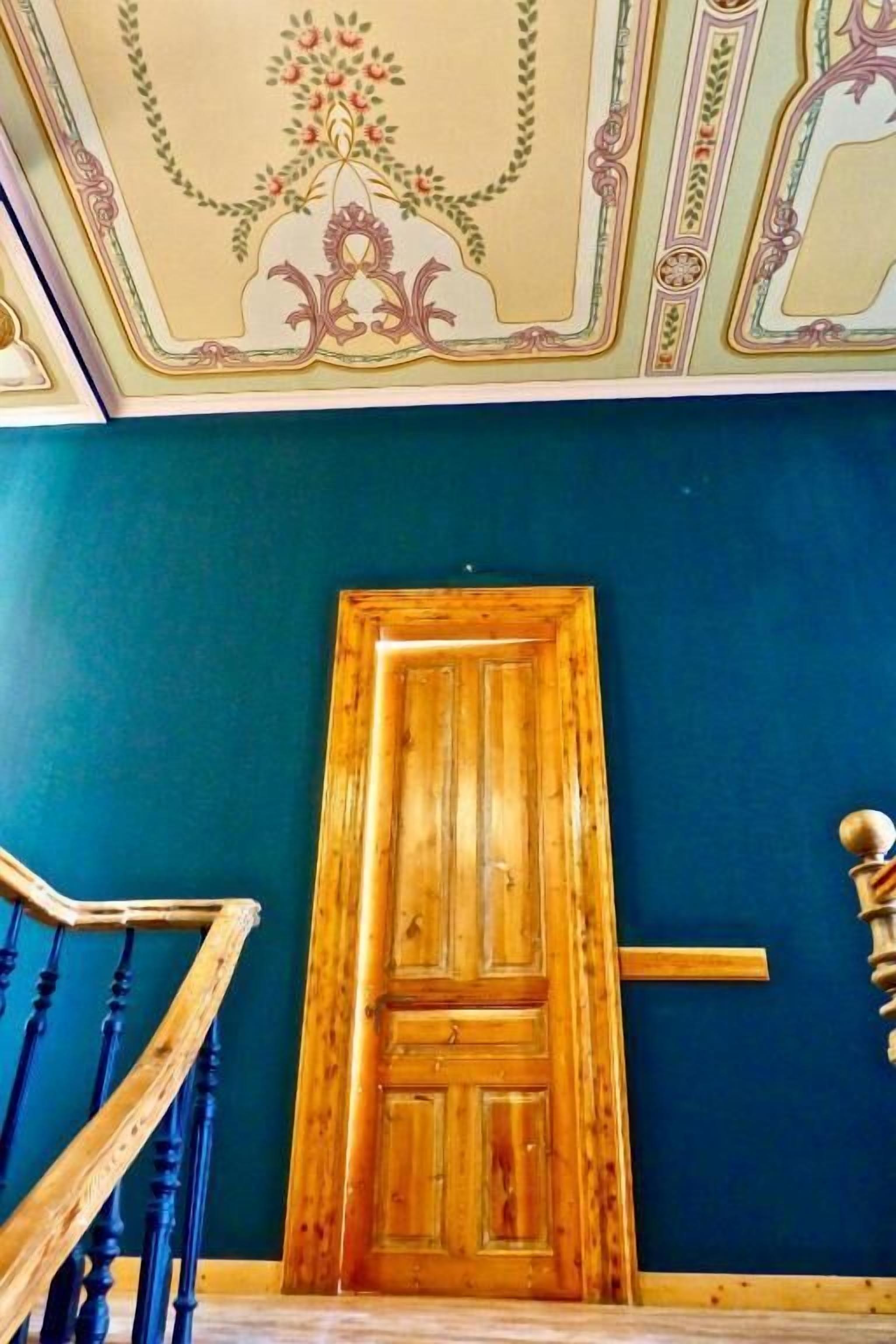 A wooden door in a room with teal walls and a ceiling with floral and geometric patterns.