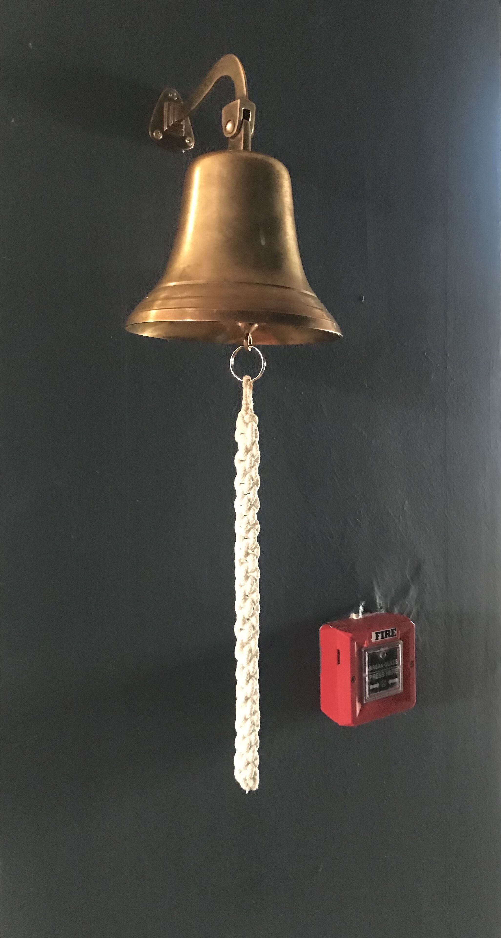 A brass bell with a white tassel hanging from a black wall.