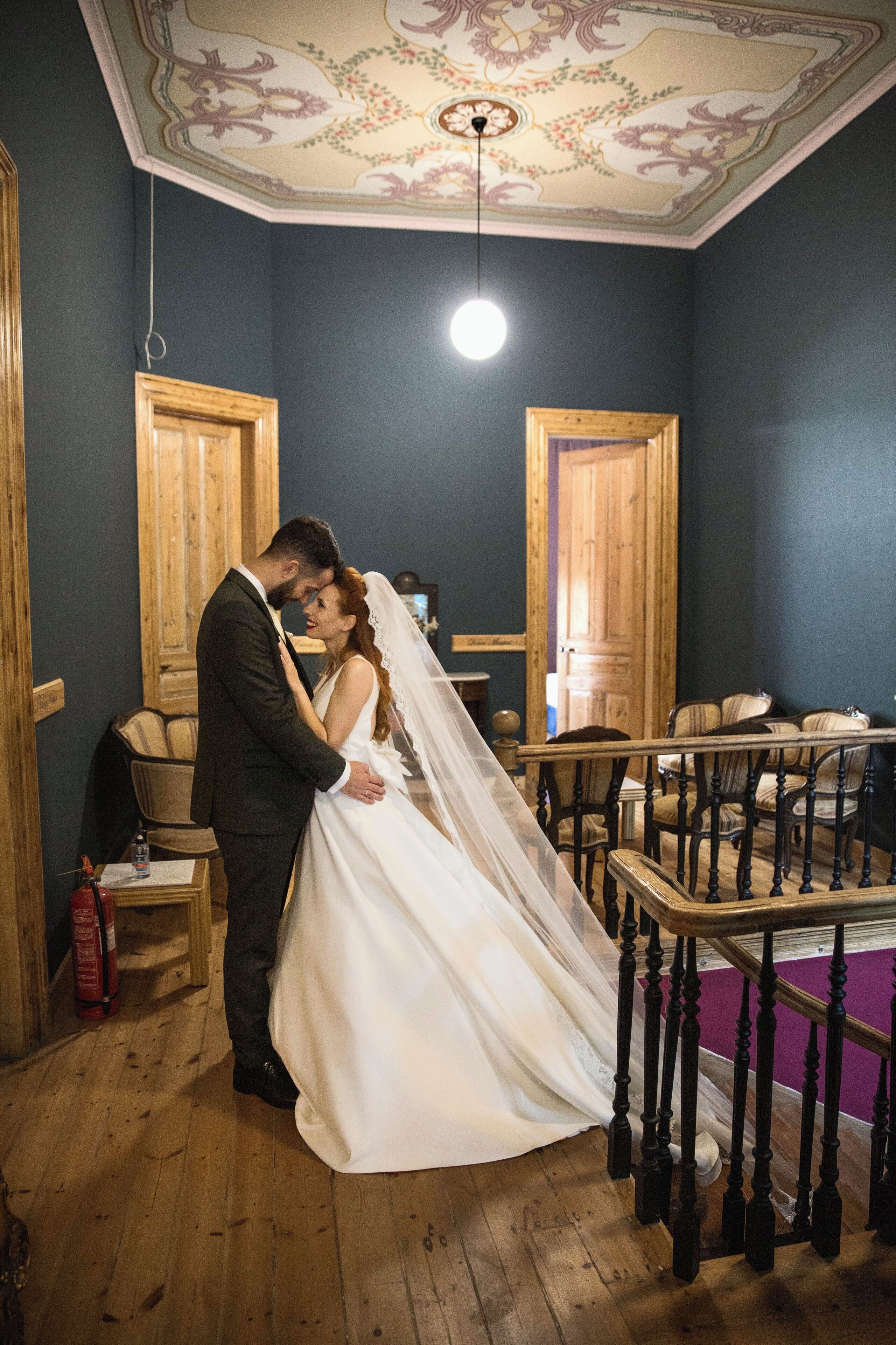 A couple embracing in a room with wooden floors, a decorative ceiling, and a staircase with wooden railings.