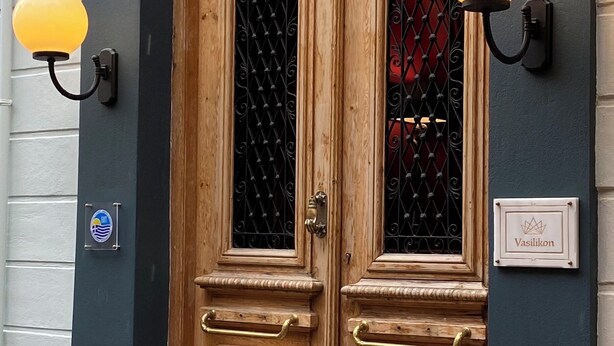 A wooden double door with a wrought iron gate, a sign above the door reading "Vasilikon Hotel," and two wall-mounted light fixtures on either side of the door. - VASILIKON Hotel 1888 the past is present (Kalamata)