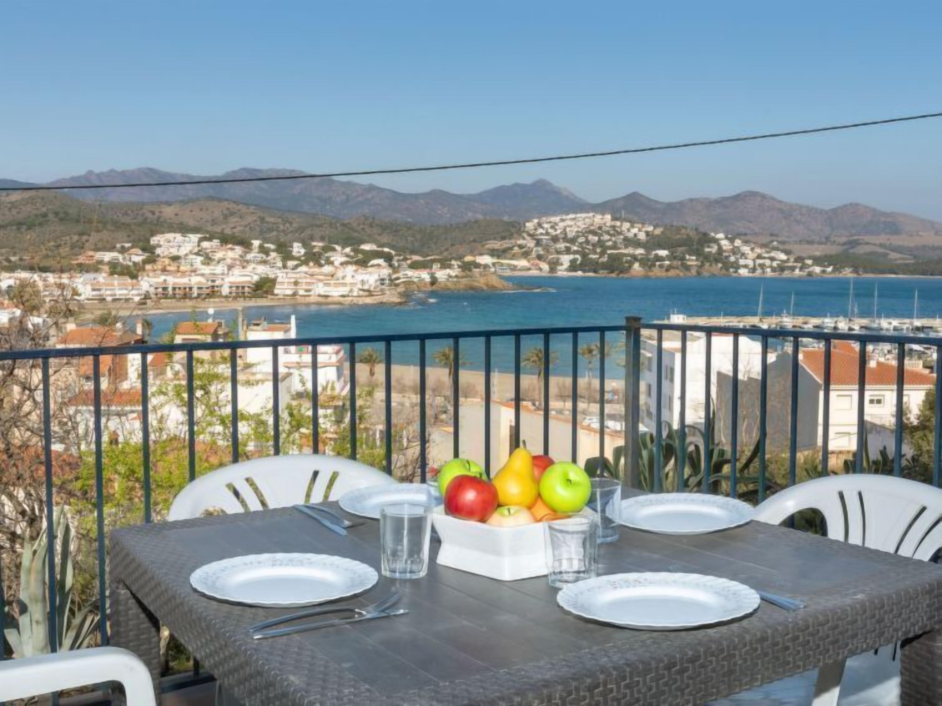 A balcony with a table set for two, overlooking a coastal town and the sea.