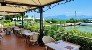 An outdoor patio with tables and chairs, overlooking a body of water and a mountainous landscape.