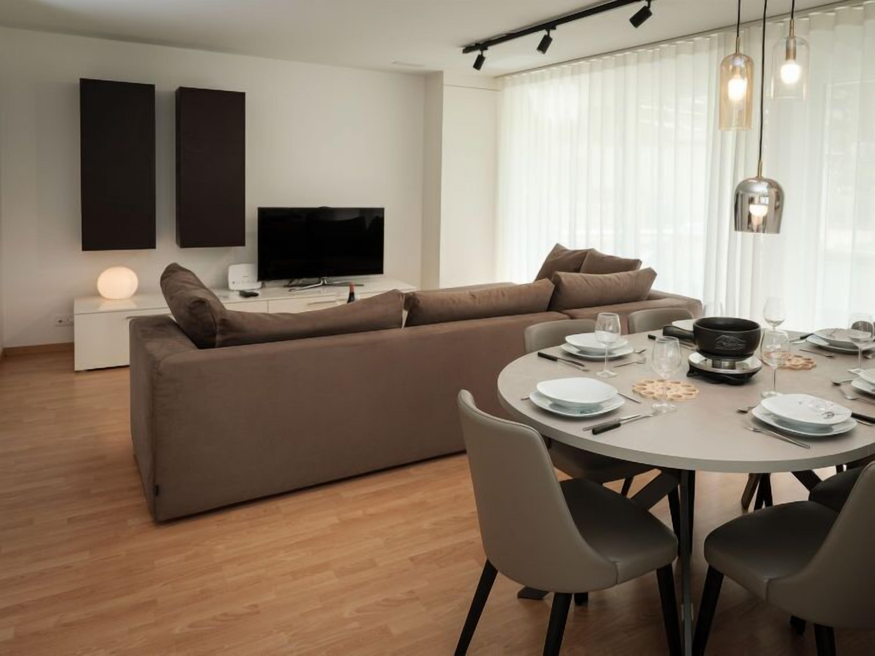 A modern living room with a sofa, a flat-screen TV, a dining table set for four, and a large window with sheer curtains.