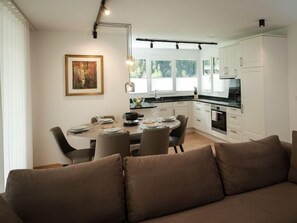 Una cocina moderna con armarios blancos, una mesa de comedor preparada para cuatro personas y un sofá en primer plano.