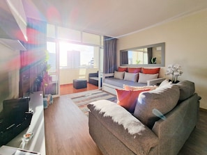 A modern living room with a sofa, a coffee table, and a TV. There is a balcony with a view of buildings and a plant.