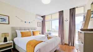 Ett hotellrum med en stor säng, ett nattduksbord, en lampa, ett fönster med gardiner och utsikt över staden.
