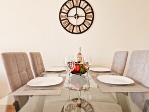 A dining table with a glass top, surrounded by cushioned chairs, a wall clock, and a fruit bowl with a wine glass. - Flat Praia Rocha Varandas Atlantis (Portimão)