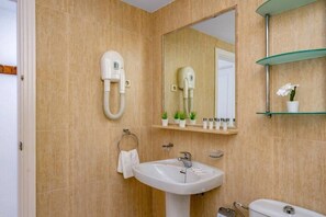 Un baño con azulejos color beige, un lavamanos blanco, un espejo, un secador de pelo y un estante con artículos de aseo.