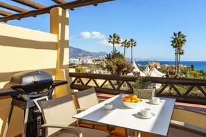 Un balcón con una mesa lista para el desayuno, con vistas a la costa, palmeras y edificios.