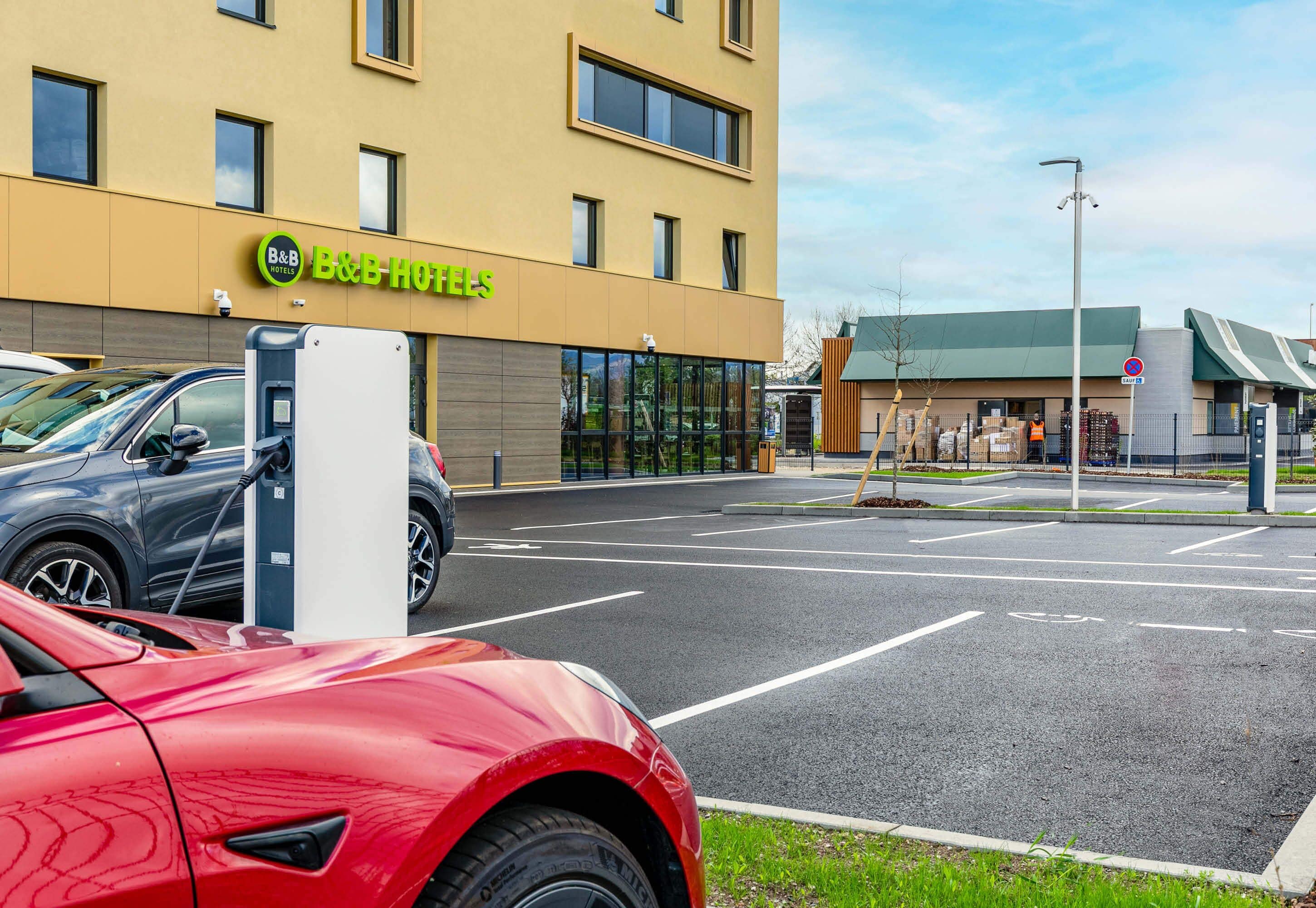 Un hotel moderno con fachada amarilla, una zona de estacionamiento con puntos de carga para vehículos eléctricos y un cielo despejado.
