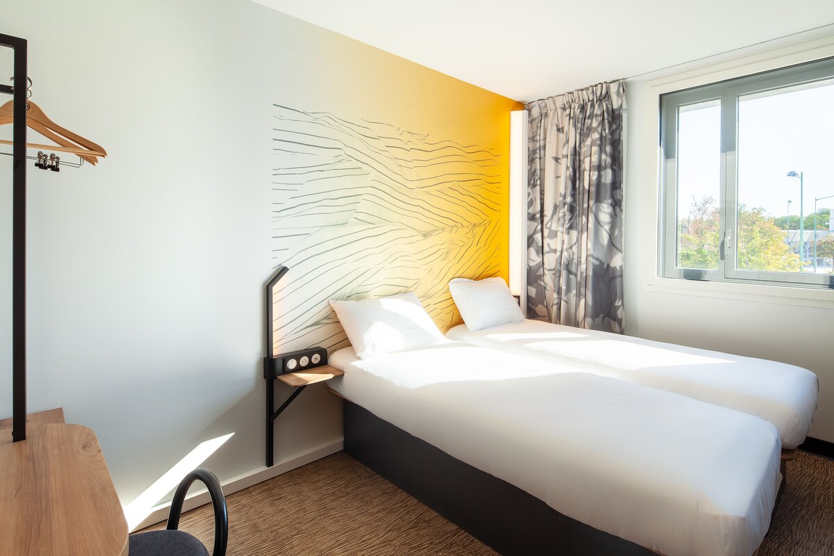 Een hotelkamer met een bed, een bureau, een stoel, een nachtkastje, een kunstwerk aan de muur en een raam met gordijnen.