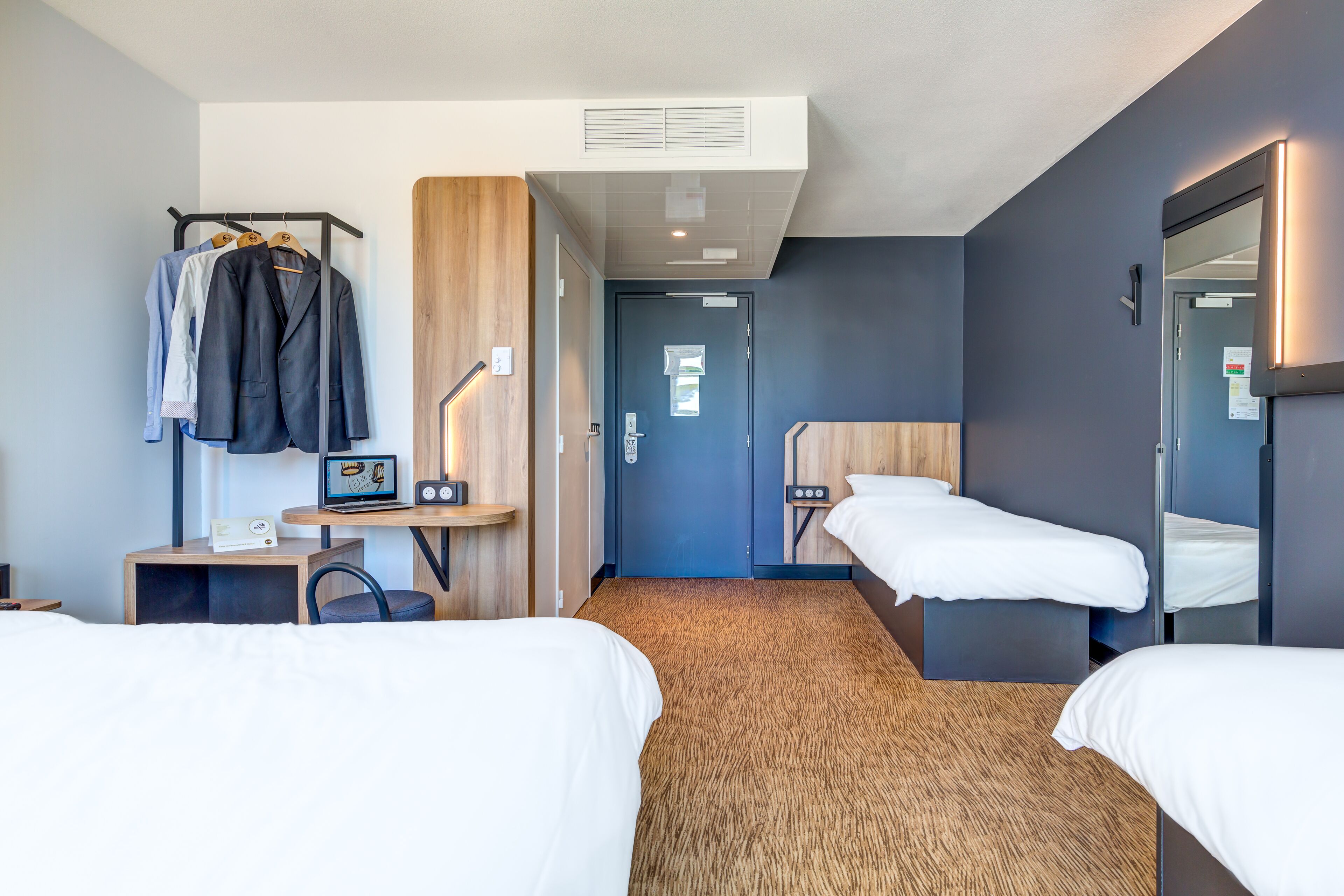 A hotel room with two single beds, a desk with a chair, a coat rack with suits, a small table with a lamp, and a large mirror.