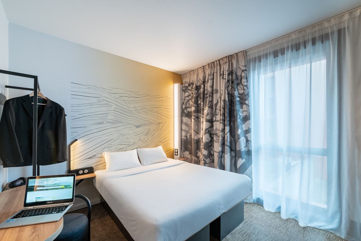 Ein modernes Hotelzimmer mit einem ordentlich bezogenen Bett, einem Laptop auf einem Schreibtisch und einem großen Fenster mit durchsichtigen Vorhängen.