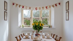 Une table à manger est dressée avec des bols, des tasses et des soucoupes blancs, entourée de chaises en bois. Une banderole portant l’inscription « PEACH COTTAGE » est suspendue au-dessus de la table.