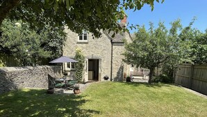 Une maison en pierre avec un mur en pierre, une cour gazonnée, une terrasse avec des meubles d’extérieur et une clôture en bois.