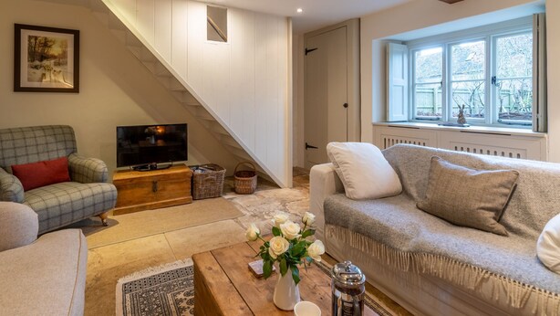 Un salon confortable avec un canap é, un fauteuil et un téléviseur posé sur un meuble en bois. Un escalier mène à une autre pièce, et une fenêtre offre une vue sur les arbres.