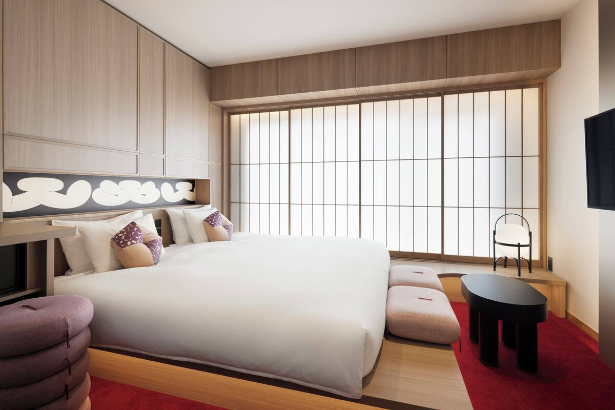 A modern hotel room with a large bed, a desk, a chair, and a view of the city through large windows.
