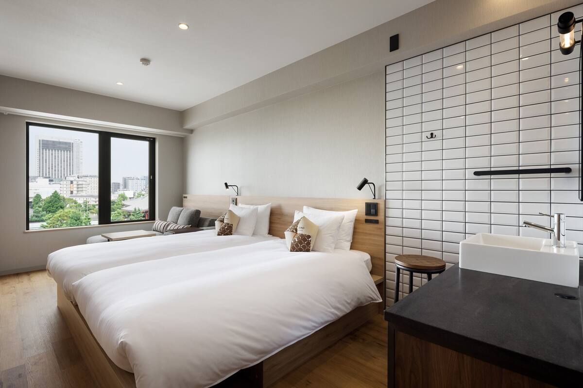 A modern hotel room with two beds, a large window, a city view, a bathroom with a sink, and a small stool.