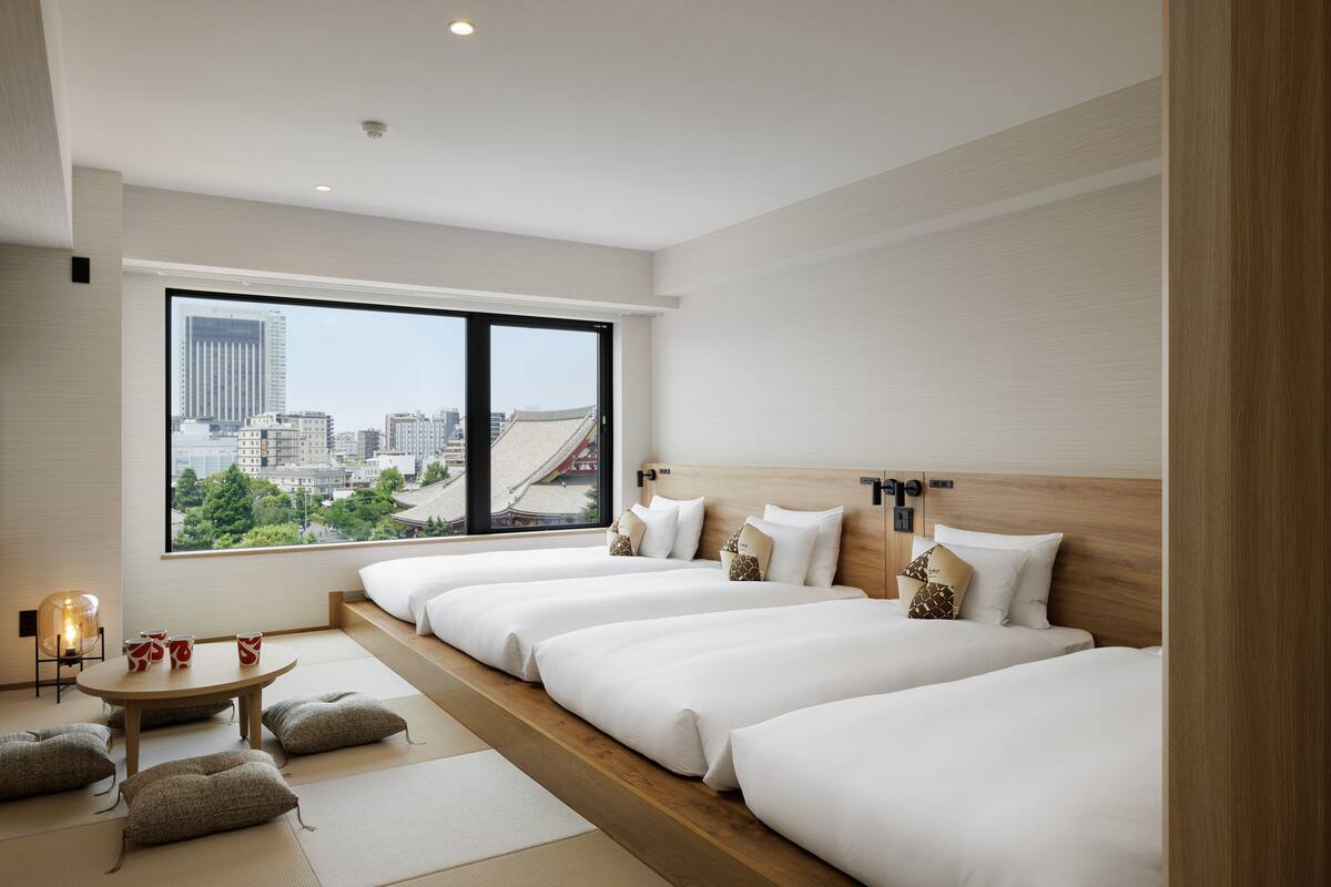 A modern hotel room with a city view, multiple beds, a small table with cups, and a traditional Japanese floor mat.