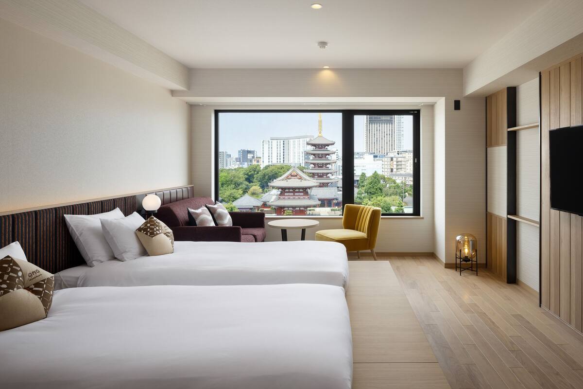 A hotel room with two beds, a sofa, a small table, and a chair. There is a large window offering a view of a traditional pagoda and modern buildings.