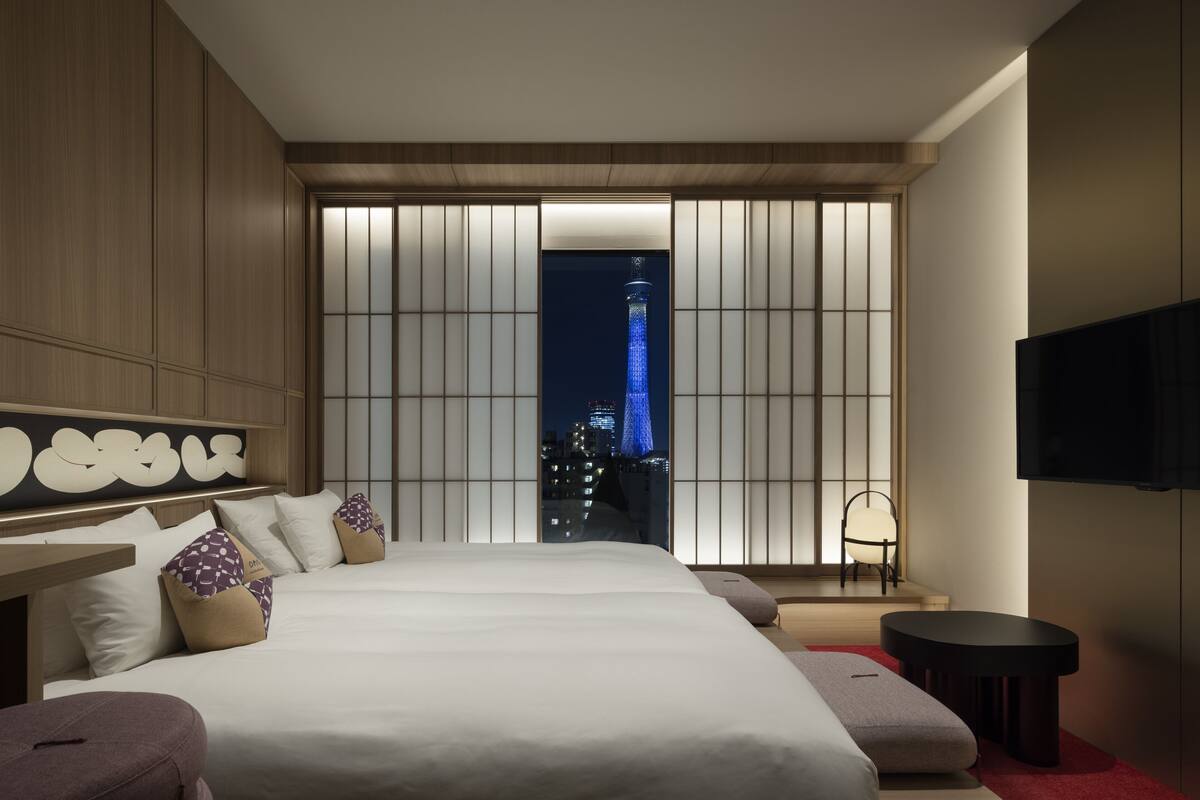 A modern hotel room with a large bed, a TV, a seating area, and a view of a cityscape at night.