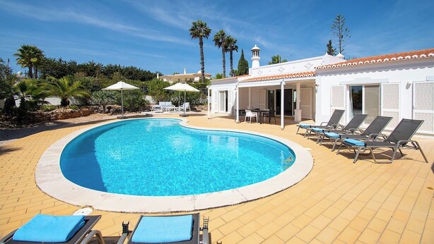 A swimming pool with lounge chairs and umbrellas, surrounded by palm trees and a building with a tiled roof. - Vivenda Pomba Beautifully renovated with heated pool! (Carvoeiro)